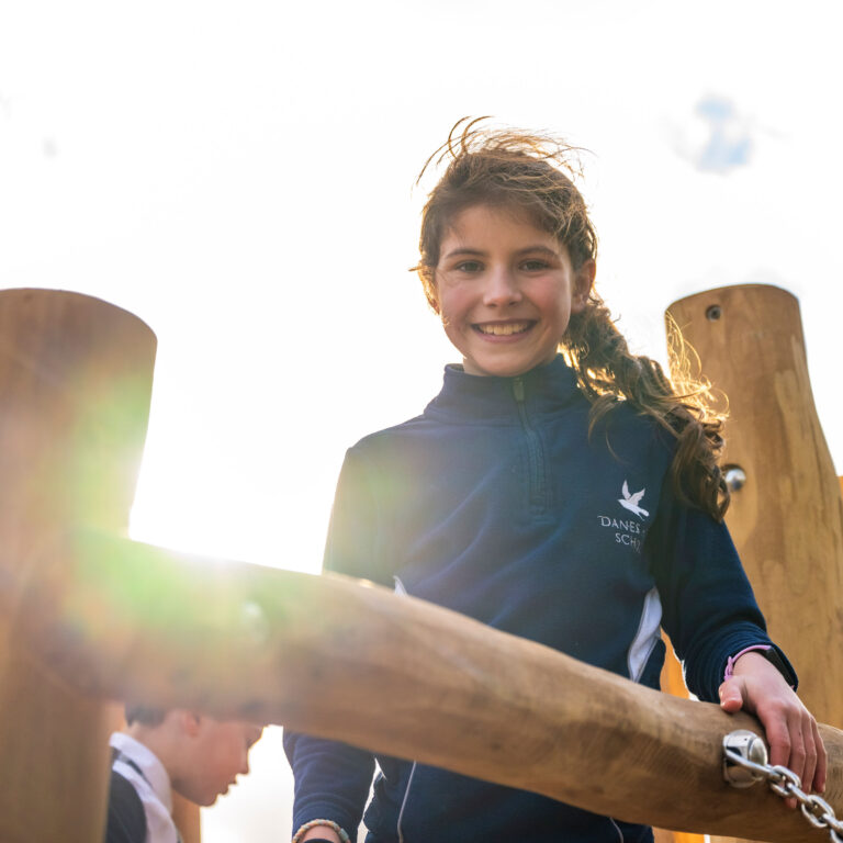 Smiling girl wearing a navy jacket on a wooden playground structure with sunlight glare behind her.